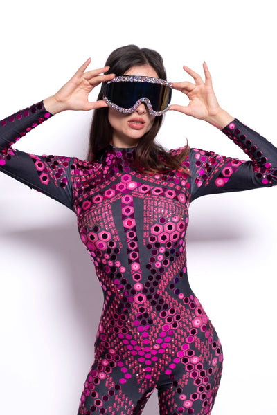 A woman wearing a form-fitting full-body bodysuit with a black base and an all-over pink and magenta circular geometric pattern, holding a pair of black ski goggles with rhinestone trim up to her eyes against a plain white background.